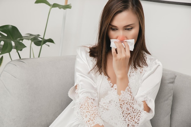 Symptoms of allergic rhinitis in women. sick woman in white nightwear with a cold blowing her nose into a tissue paper at home. cold weather allergies