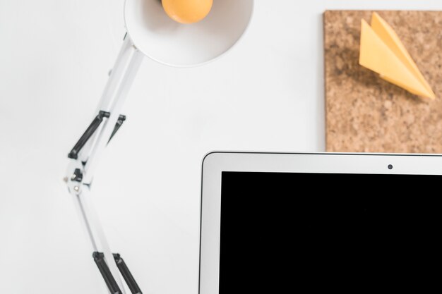 Table lamp, laptop and yellow paper plane on cork board against white background