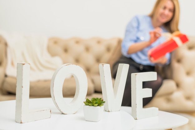 Table with love sign near young lady with gift box on sofa