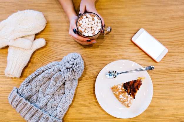 Table with white winter gloves, piece of pie, knitted hat, phone and hot chocolate in hands of girl.