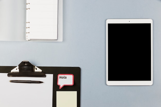 Tablet near notebook, clipboard with paper and frame with Hola inscription