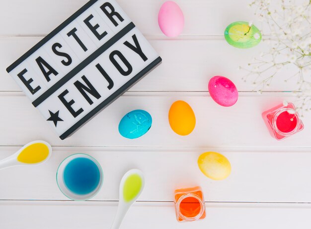 Tablet with Easter enjoy title near eggs between flower twig and cans with dye liquid