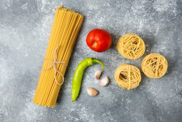 Tagliatelle raw pasta nests, spaghetti and vegetables on marble surface. 