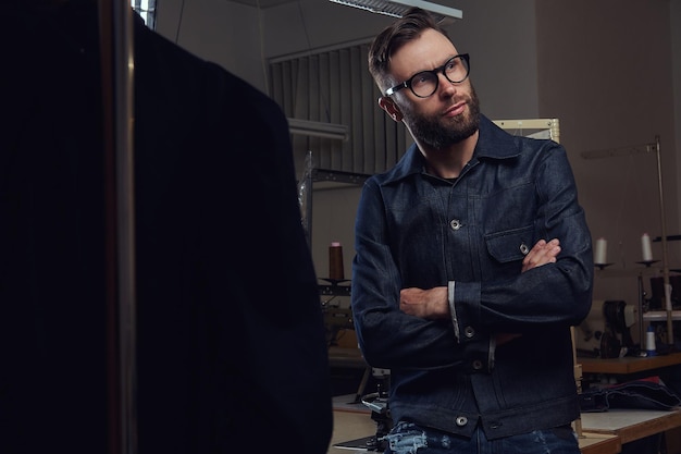 Free photo tailor standing near a table with sewing machine and looking away at a sewing workshop.