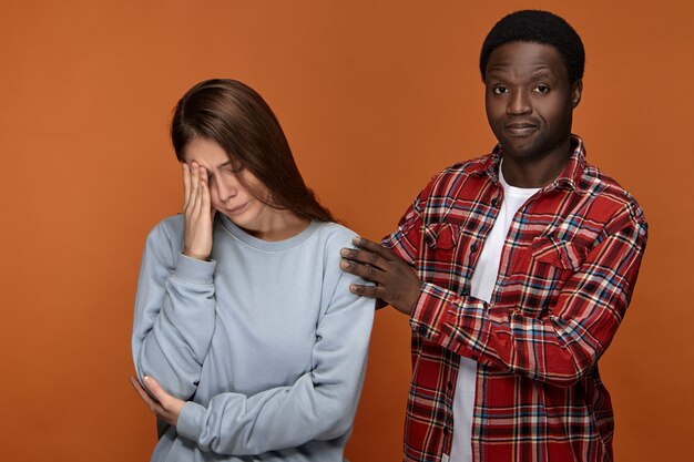 Take it easy. Attractive stylish African American guy hugging his upset depressed European girlfriend who is feeling unwell because of terrible headache, touching her head with eyes closed