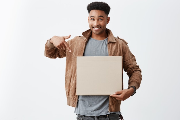 Free photo take a look at this. close up portrait of young beautiful black-skinned male university student with afro hairstyle in stylish autumn clothing pointing at carton board in hands with happy expression