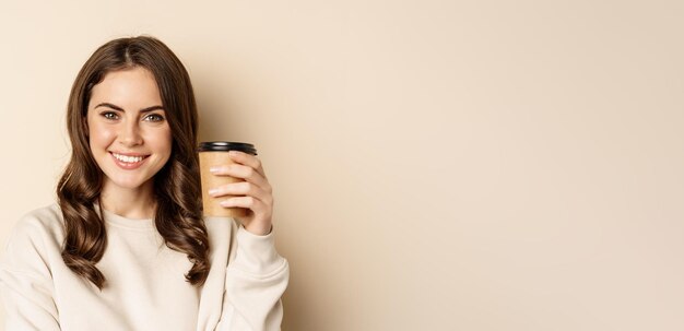 テイクアウトとカフェのコンセプト美しいフェミニンな女性が笑顔を浮かべて、beig に対してポーズをとるコーヒーのカップを保持しています