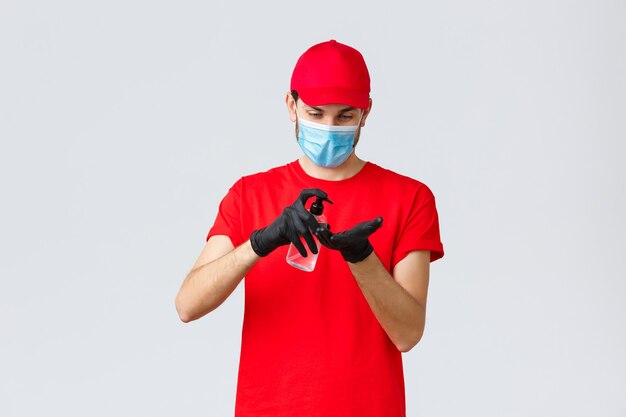 Еда на вынос, доставка еды и продуктов, концепция бесконтактных заказов covid-19. Курьер или сотрудник в красной футболке и форме кепки, в маске и резиновых перчатках, нанесите дезинфицирующее средство для рук.