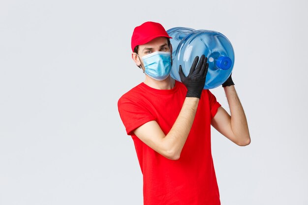 Takeaway, food and groceries delivery, covid-19 contactless orders concept. Young courier in red uniform, cap and face mask with gloves, delivering bottled water to your office or home