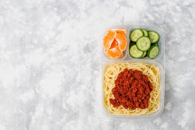 Takeaway spaghetti bolognaise in a plastic lunch box and fruit slice on light 
