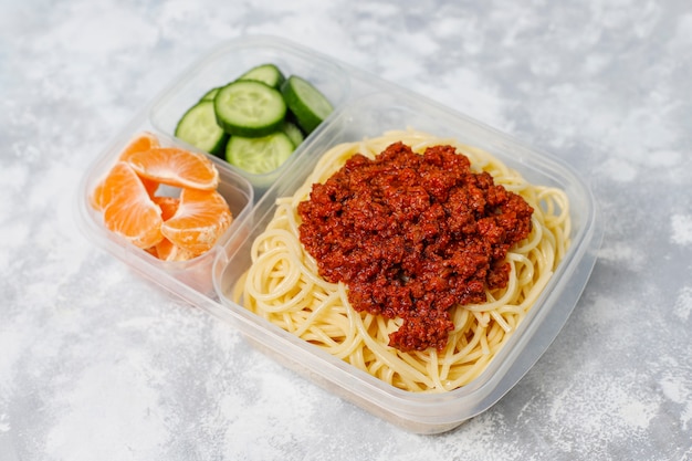 Takeaway spaghetti bolognaise in a plastic lunch box with detox drink and fruit slice on light 