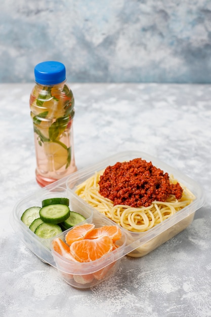 Takeaway spaghetti bolognaise in a plastic lunch box with detox drink and fruit slice on light 