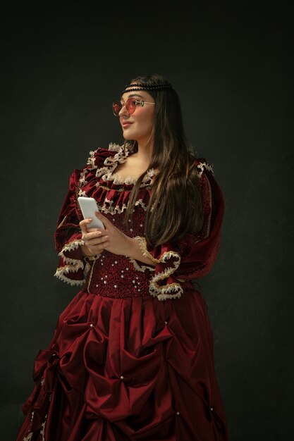 Takes selfie wearing modern eyewear. Medieval young woman in red vintage clothing on dark background.