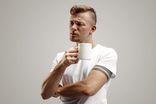 コーヒーブレイク。灰色のスタジオに立っている間コーヒーカップを保持しているハンサムな若い男