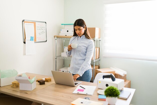 Taking a little break from office work. Beautiful business owner drinking a cup of coffee in the morning while looking at her laptop