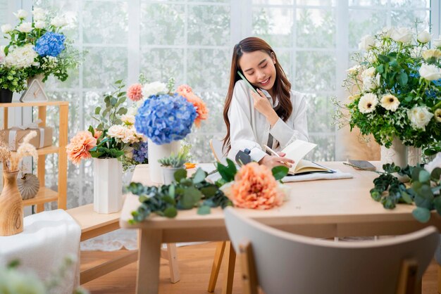 Принимающий заказ флорист, работающий домамолодой азиатский флорист составляет список от клиента, чтобы организовать доставку вазы букета цветов Магазин цветочного дизайна счастье улыбающаяся юная леди делает вазу для цветов