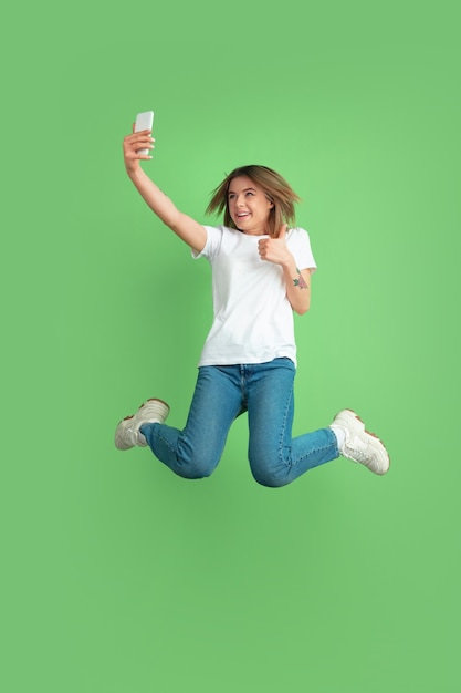 Taking selfie in jump. Caucasian young woman's portrait isolated on green studio wall