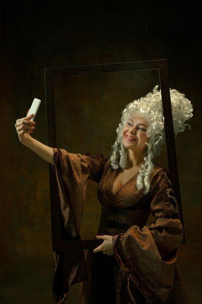 Taking selfie. Portrait of medieval young woman in vintage clothing with wooden frame on dark background. Female model as a duchess, royal person. Concept of comparison of eras, fashion, beauty.