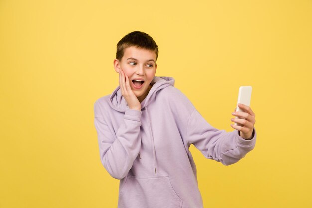 Taking selfie, vlog. Caucasian girl's portrait isolated on yellow background with copyspace. Beautiful female model in hoodie. Concept of human emotions, facial expression, sales, ad, fashion.