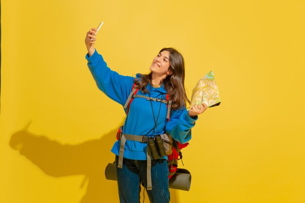 Taking selfie or vlog. Portrait of a cheerful young caucasian tourist girl with bag and binoculars isolated on yellow studio background. Preparing for traveling. Resort, human emotions, vacation.