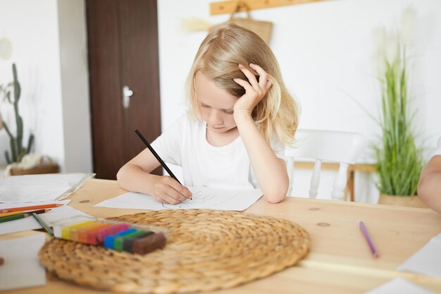 家で楽しい時間を過ごし、頭を手に置いてテーブルに座って、絵を描いたり、スケッチしたり、黒鉛筆を使ったりする才能のある金髪のヨーロッパの少年。木製の机に集中した男子生徒の着色