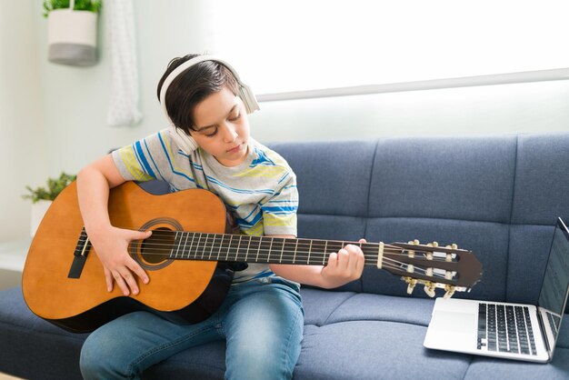 Талантливый мальчик слушает песню в наушниках, играя на акустической гитаре и следуя видеоуроку на своем ноутбуке.