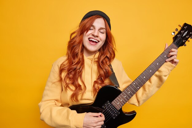 재능있는 여성 뮤지션이 일렉트릭 기타를 부르며 좋아하는 노래를 부르며 무대에서 공연을 준비하고 모자를 쓰고 스웨트 셔츠에 긴 빨간 머리를 가짐