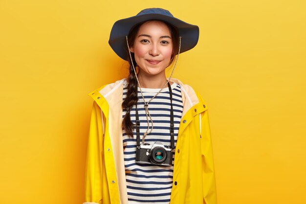 Talented female photographer makes professional shots during adventure trip, uses retro camera, wears stylish hat, yellow raincoat, enjoys holidays