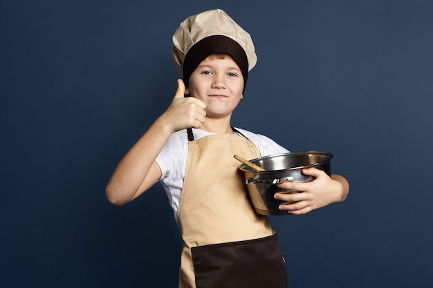 Talented little boy chef in cap and apron holding big metal saucepan, smiling confidently, showing thumbs up gesture while cooking delicious meal. Food, cuisine, cookery and gastronomy concept