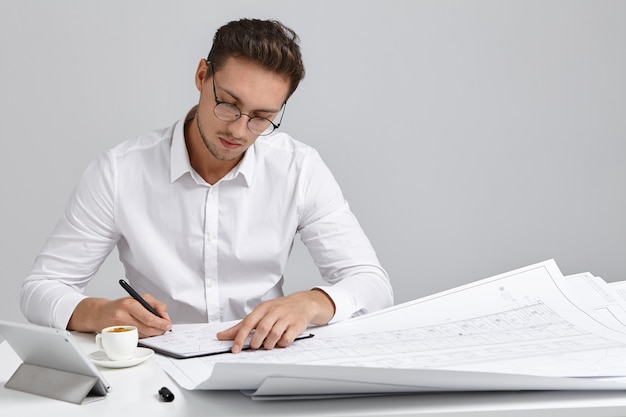 Talented young European bearded chief engineer wearing round glasses and white formal shirt sitting at his workplace