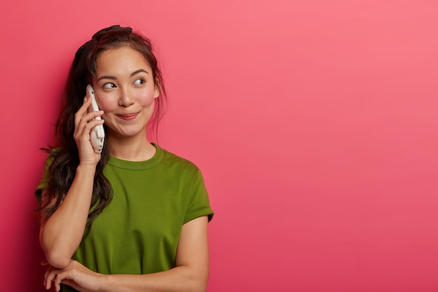 Talkative teenage girl spends hours talking via smartphone, holds cellphone close to ear, calls friends, enjoys nice conversation, looks with thoughtful expression aside, dressed in casual wear