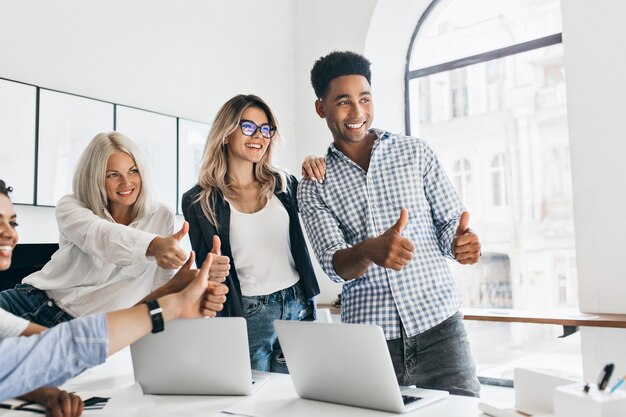 Tall african manager with big smile holding thumbs up and looking away. Charming girls working as managers posing in office next to table with laptops on it and laughing.