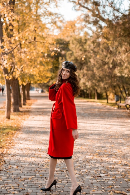 暖かい赤いコート秋の流行のファッション、ストリートスタイル、ベレー帽の帽子をかぶって公園を歩いている巻き毛の背の高い魅力的なスタイリッシュな笑顔の細い女性