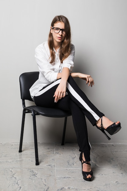 Tall brunette girl in glasses dressed up in white t-shirt and black pants sits on office chair in front of white background