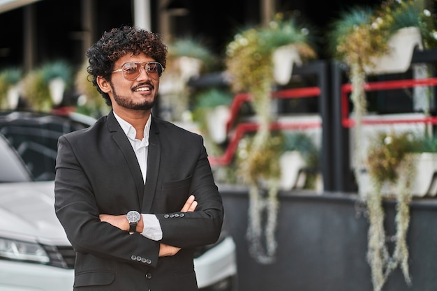 Tall Indian businessman with glasses near the car in a black suit.