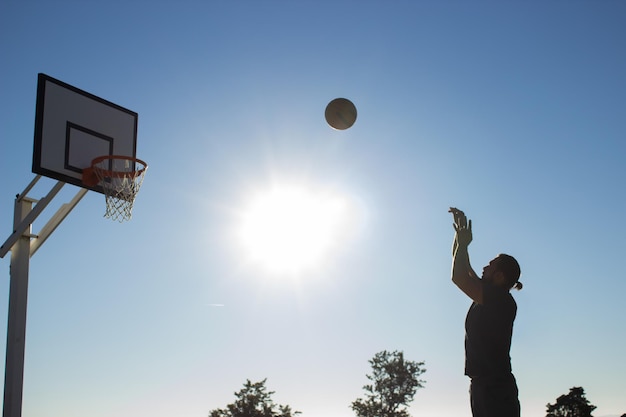 Tall man playing basketball alone on sunny day outdoors at sports ground. Man throwing basketball into basket against zenith sky background. Active lifestyle, sport, motivation concept