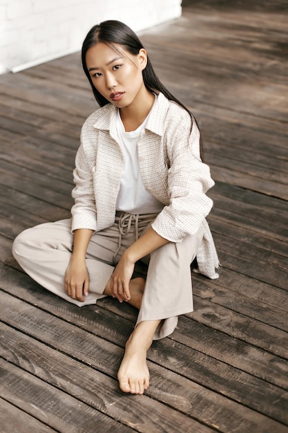 Tanned Asian woman in beige pants and plaid jacket sits on wooden floor Portrait of brunette longhaired lady in stylish outfit looks into camera