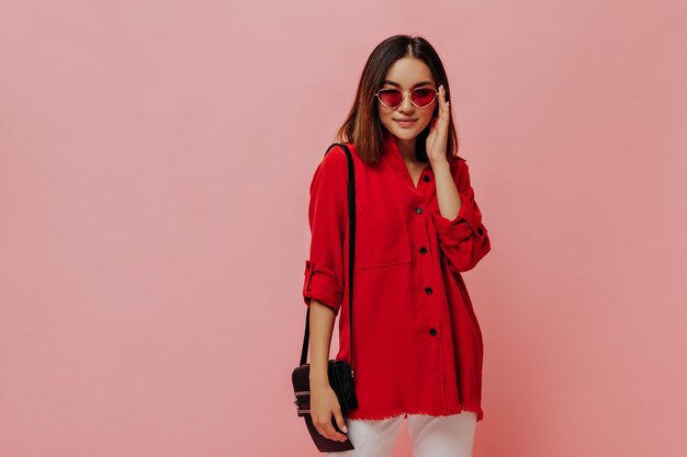 特大の赤いシャツの日焼けしたアジアの女性はサングラスをかけ、ピンクの背景にハンドバッグでポーズをとる