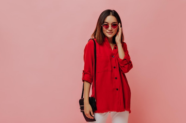 特大の赤いシャツの日焼けしたアジアの女性はサングラスをかけ、ピンクの背景にハンドバッグでポーズをとる