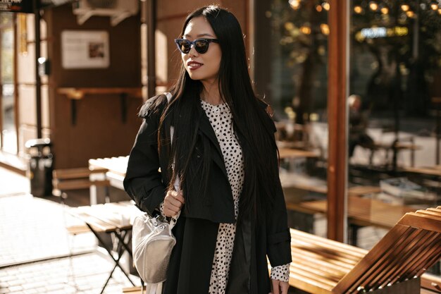 Tanned Asian woman in stylish trench coat and white dress walks outside Pretty brunette lady in sunglasses smiles