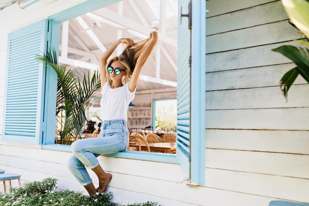Tanned barefooted woman in vintage jeans playing with her beautiful long hair Photo of fashionable girl in sparkle sunglasses chilling on window sill