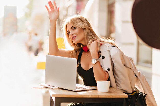 Tanned blonde girl in beige coat with leather bag saw friend and waving hand to him. Ecstatic female model with laptop having fun in cafe during business lunch in good day.
