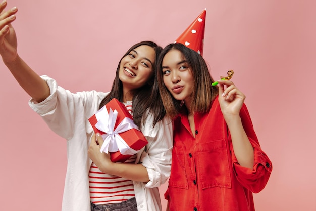 Tanned brunette Asian women take selfie and celebrate birthday on pink background Cute girl in white shirt poses with red gift box Charming lady in party hat holds party horn