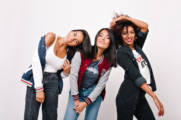 Tanned brunette girl in black denim pants posing beside cheerful sister and her african female friend. Indoor portrait of ecstatic university mates with different ethnicity having fun together.