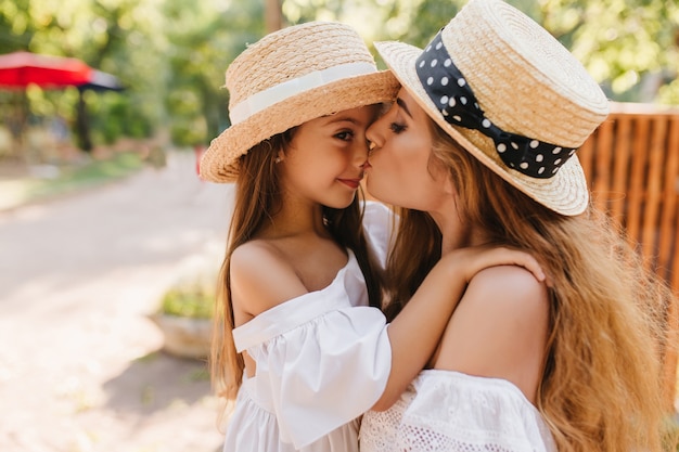 Tanned brunette girl in straw hat decorated with white ribbon embracing mom and looking away. Long-haired young woman standing beside wooden fence kissing her little daughter with love.