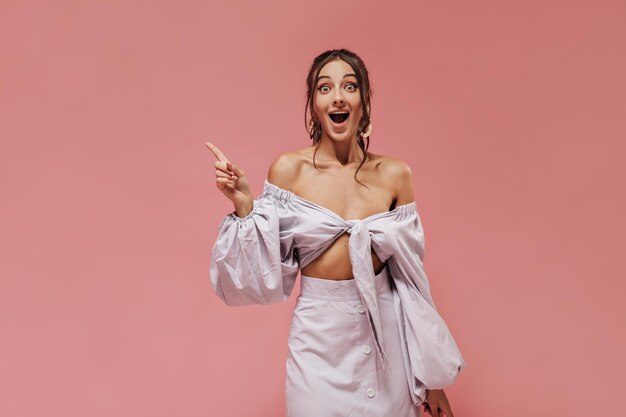 Tanned brunette in gold earrings striped cool shirt and modern skirt looking into camera and having idea on pink backdrop