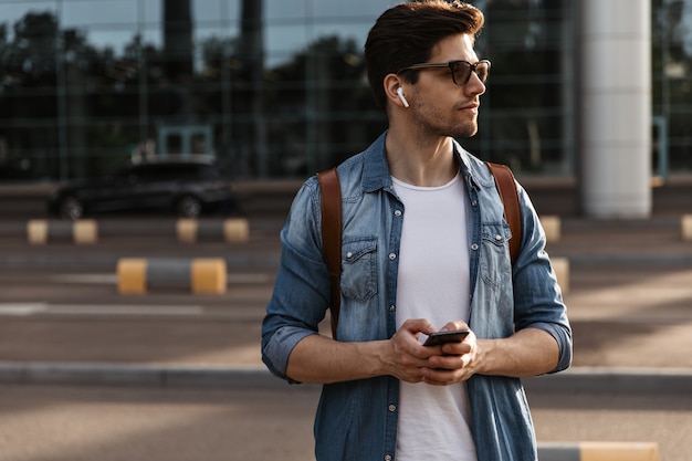 Tanned brunette man in white t-shirt, denim jacket and sunglasses poses outside