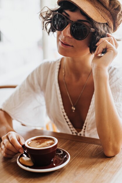 茶色の帽子とサングラスで日焼けしたブルネットの女性は、コーヒーとカフェで休んで茶色の磁器のカップを保持します