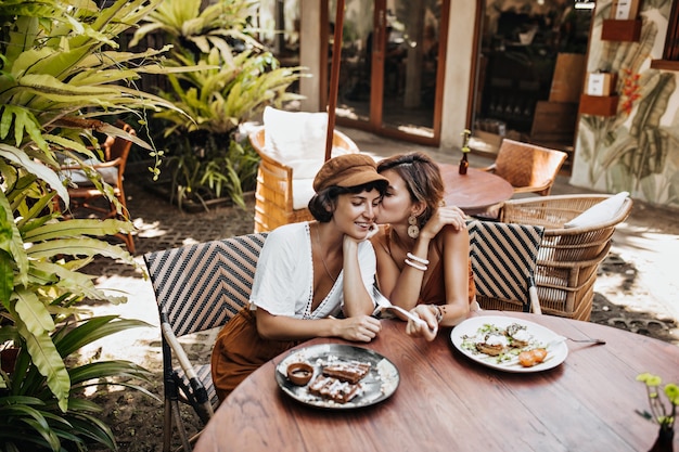 Tanned cheerful women in stylish summer outfits gossip and enjoy tasty food in street cafe