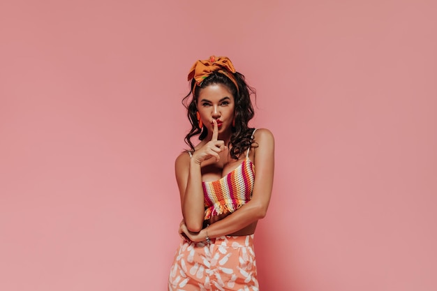 Tanned cool brunette with modern bright bandana and accessories in stylish colorful outfit looking into camera on pink background